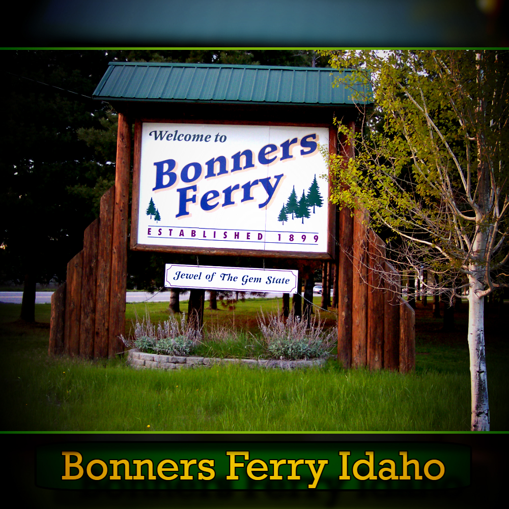A wooden sign welcomes visitors to Bonners Ferry, Idaho, established in 1893, with the tagline "Jewel of The Gem State." Amidst trees and greenery, a nearby tow truck adds a touch of modern utility to this picturesque scene.