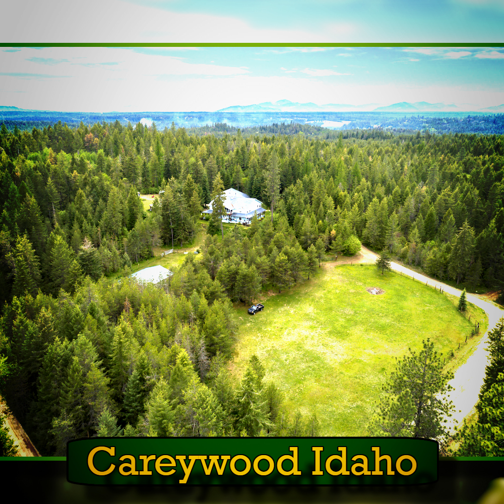 Aerial view of dense forest with a clearing featuring a house, a few buildings, and a tow truck in Careywood, Idaho.