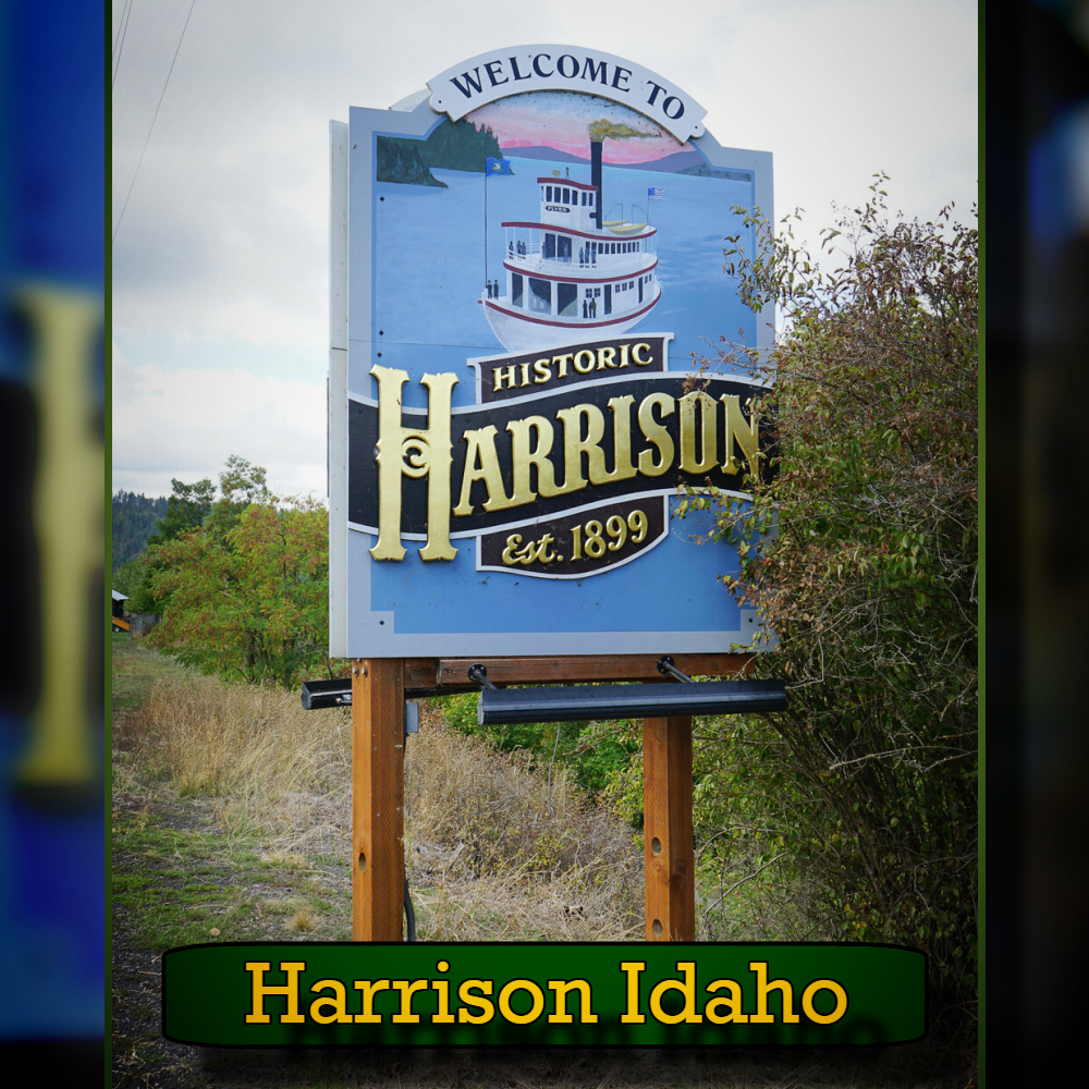 Sign reading "Welcome to Historic Harrison Est. 1899" by the roadside, with foliage around. The sign features an image of a boat on a lake and a tow truck, symbolizing the local towing services available in town.