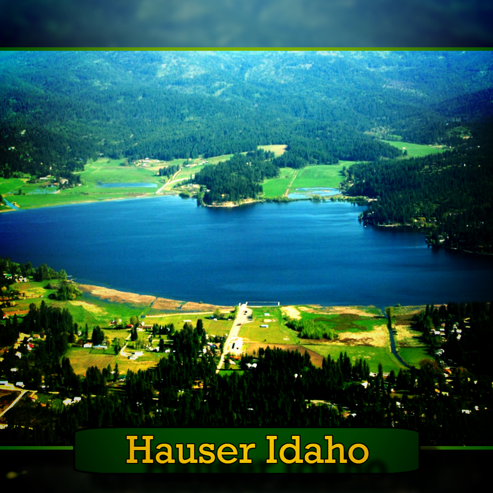 Aerial view of Hauser Lake surrounded by lush forests and fields in Hauser, Idaho, with mountains in the background. A tow truck can be seen on the road below. Text at the bottom reads "Hauser Idaho".