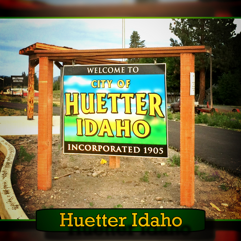 A wooden sign reads, "Welcome to City of Huetter Idaho, Incorporated 1905," situated in an outdoor area with trees in the background, with a tow truck passing by on the nearby road.