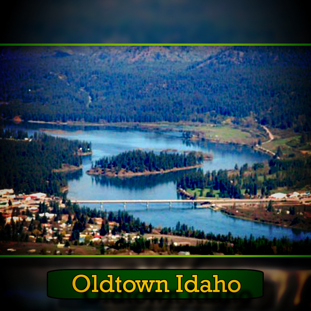 Aerial view of Oldtown, Idaho, showing a lake surrounded by forested hills and a bridge connecting parts of the town, with a tow truck making its way across the bridge.