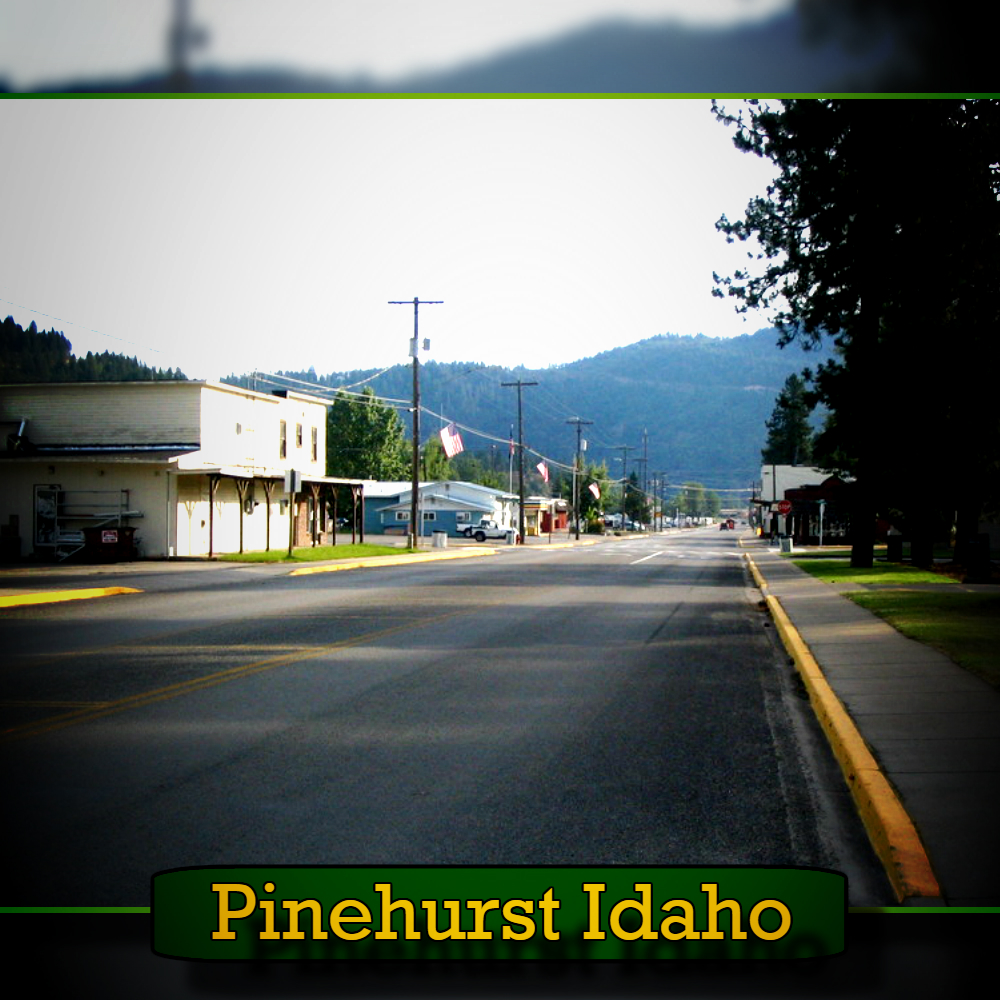 A quiet street lined with buildings and trees in Pinehurst, Idaho, showcases a mountainous background. A sign in the foreground reads "Pinehurst Idaho," and a tow truck is parked nearby, hinting at reliable local tow service available to residents and visitors alike.