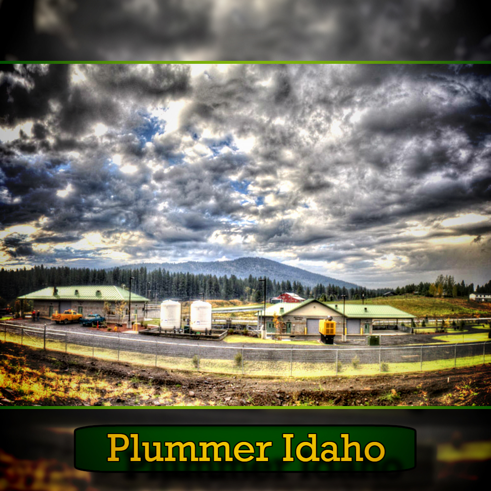 A scenic view of an industrial facility with large storage tanks and green-roofed buildings against a backdrop of a cloudy sky and forested hills, along with a tow truck in the foreground, captioned "Plummer Idaho.