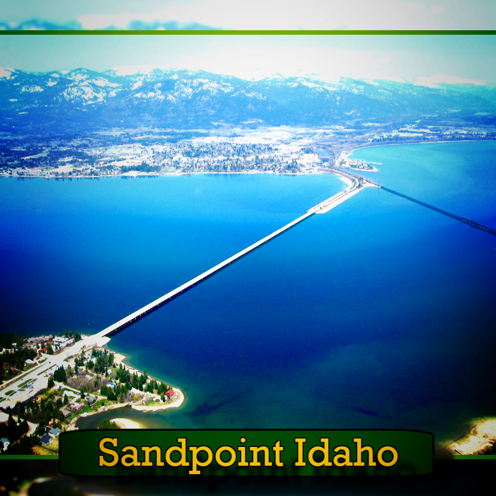 Aerial view of Sandpoint, Idaho, featuring a long bridge crossing over a large body of water with mountains in the background. A tow truck can be spotted near the bridge. The caption reads "Sandpoint Idaho.